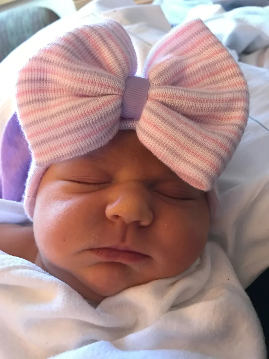 A newborn asleep in a pink and white striped hospital hat with a large bow