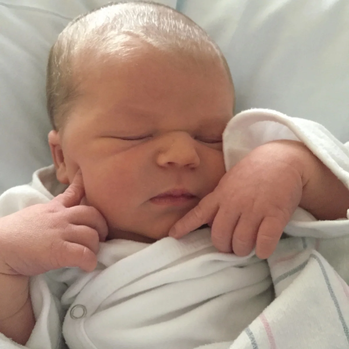 A newborn swaddled in white, both hands tucked up beside their face, fast asleep