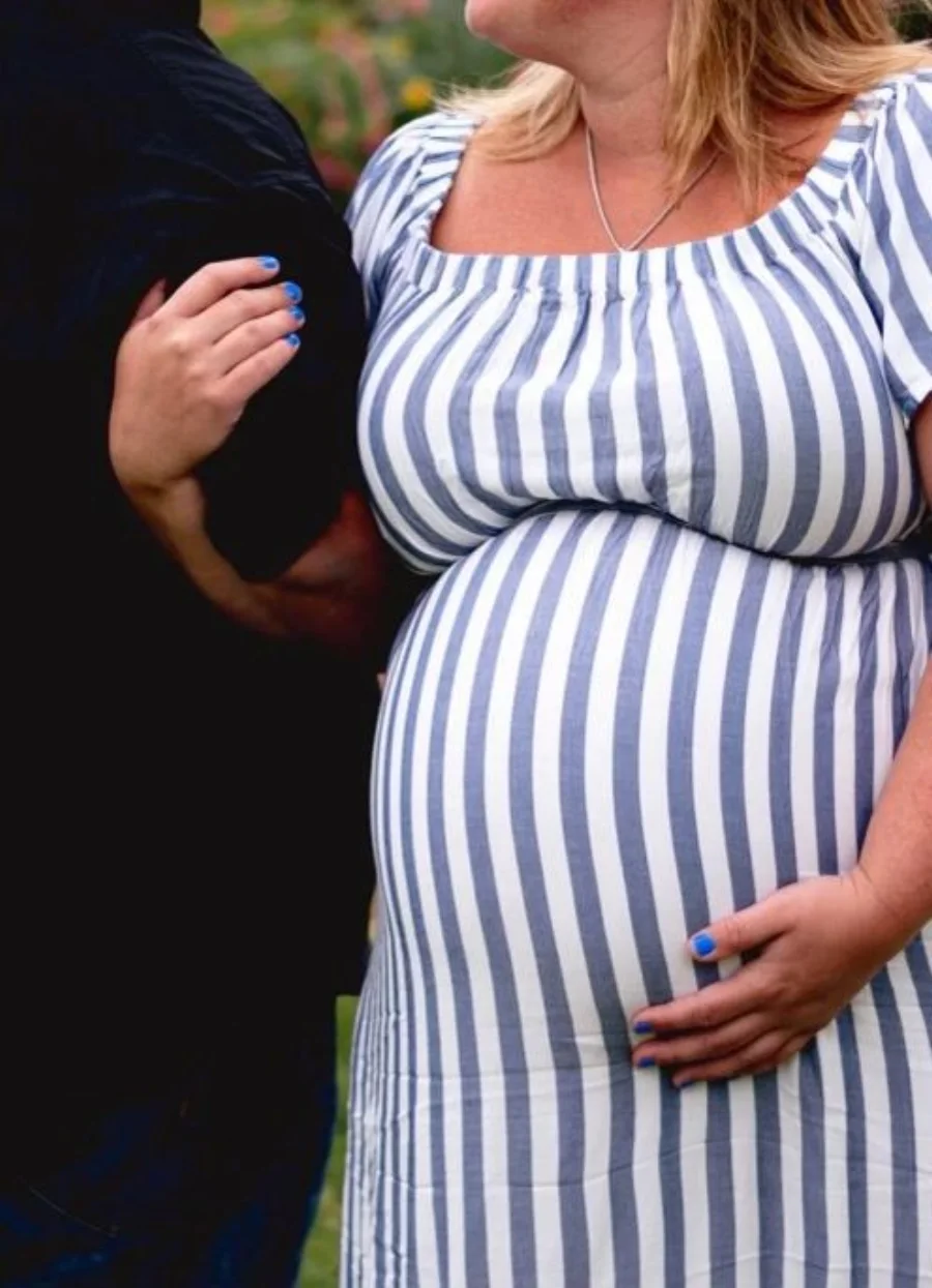 An expectant mother in a striped dress, one hand resting on her bump, holding her partner’s arm