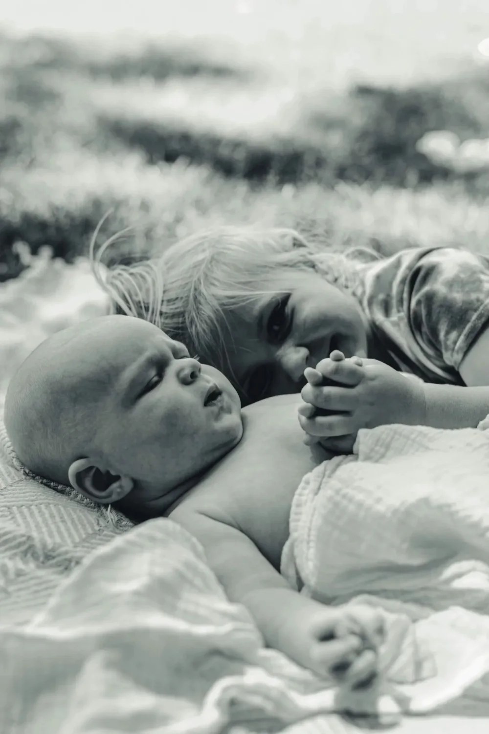 A newborn lying on a blanket outdoors while their older sibling leans in beside them, holding hands